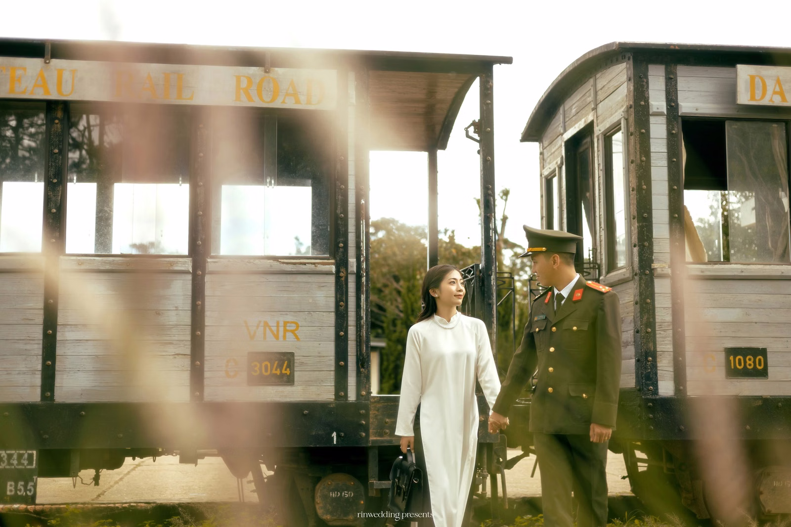 The couple stands beside a train carriage in sunshine at Ga Đà Lạt.