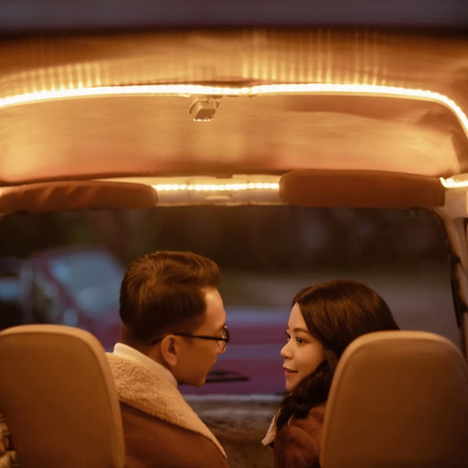 The couple sits inside a car and turns toward each other under warm yellow light.
