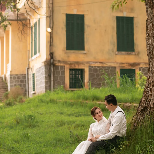 The bride and groom sit beneath a tree before a yellow mansion.