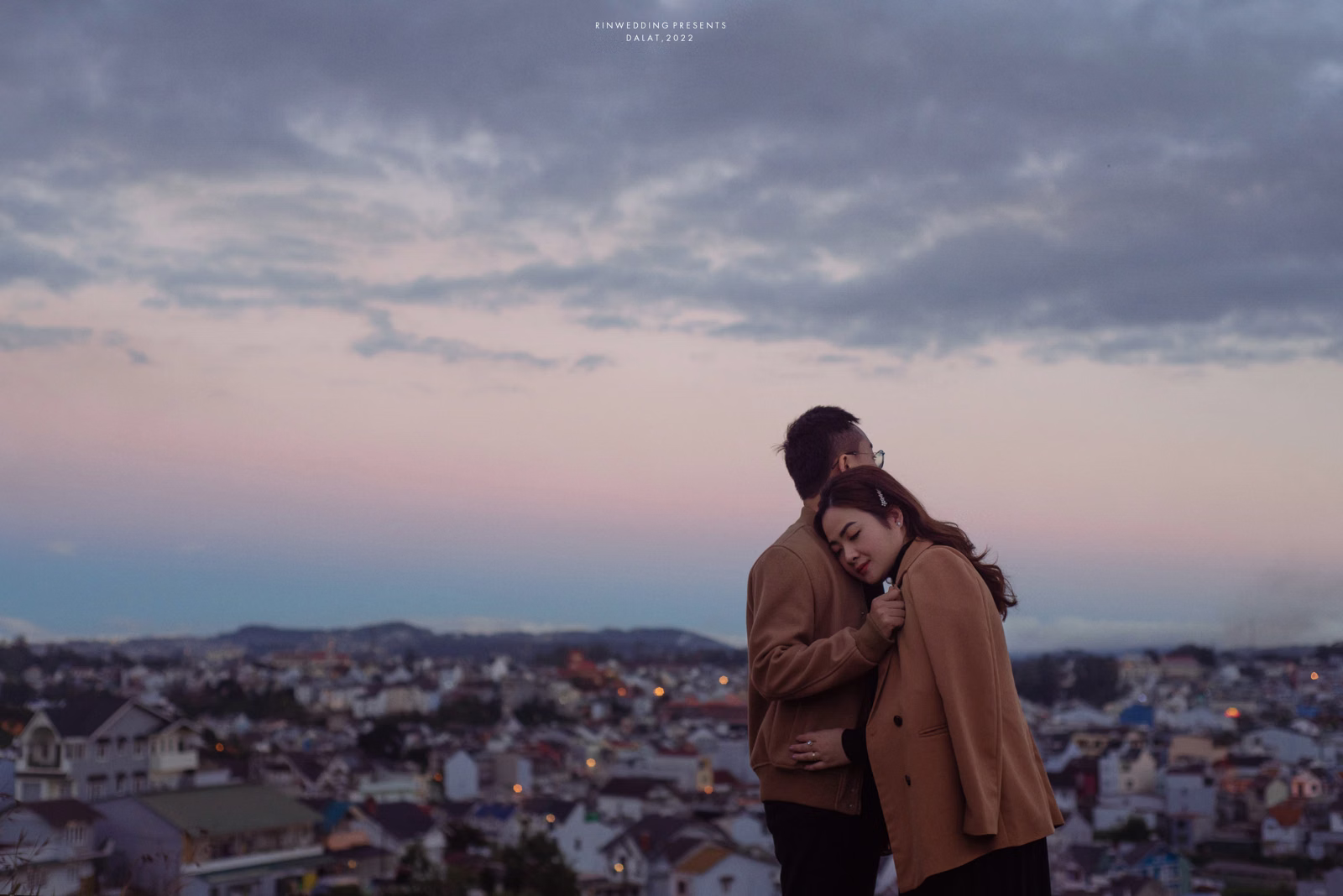 The bride rests her head on the groom's shoulder before the city at dusk.