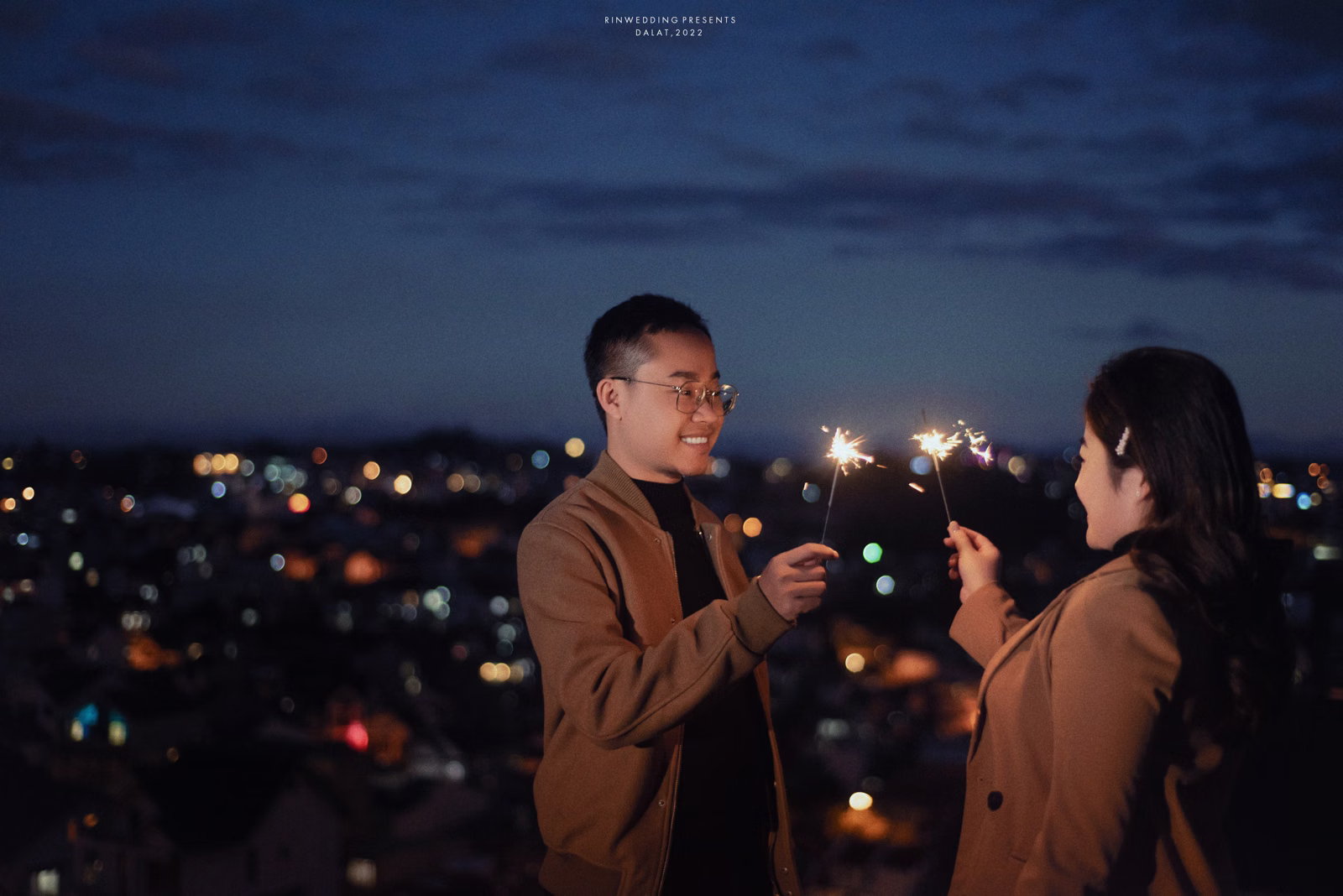 The couple holding sparklers and looking at each other against city lights at night.