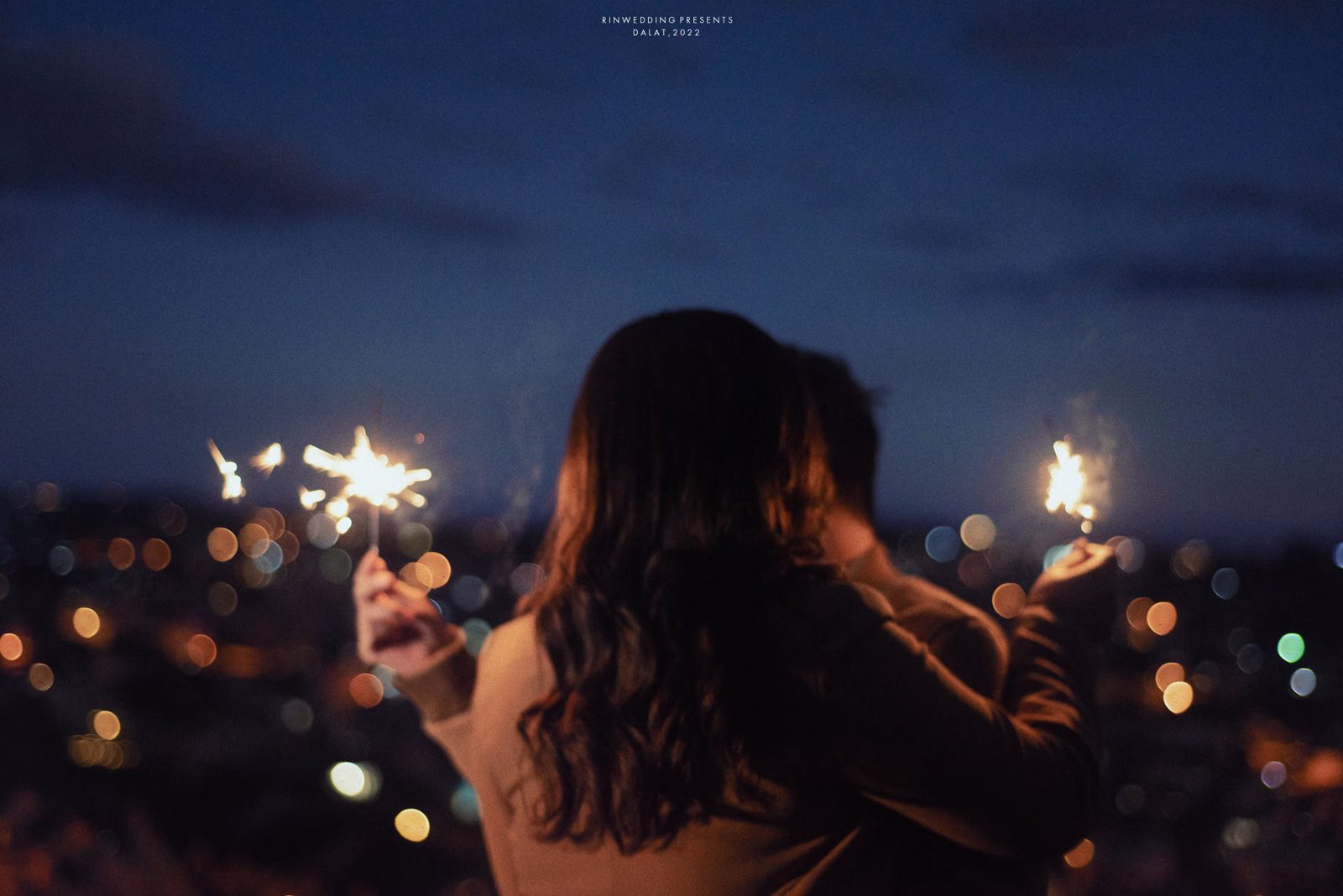 The bride seen from behind holding two sparklers before city lights.