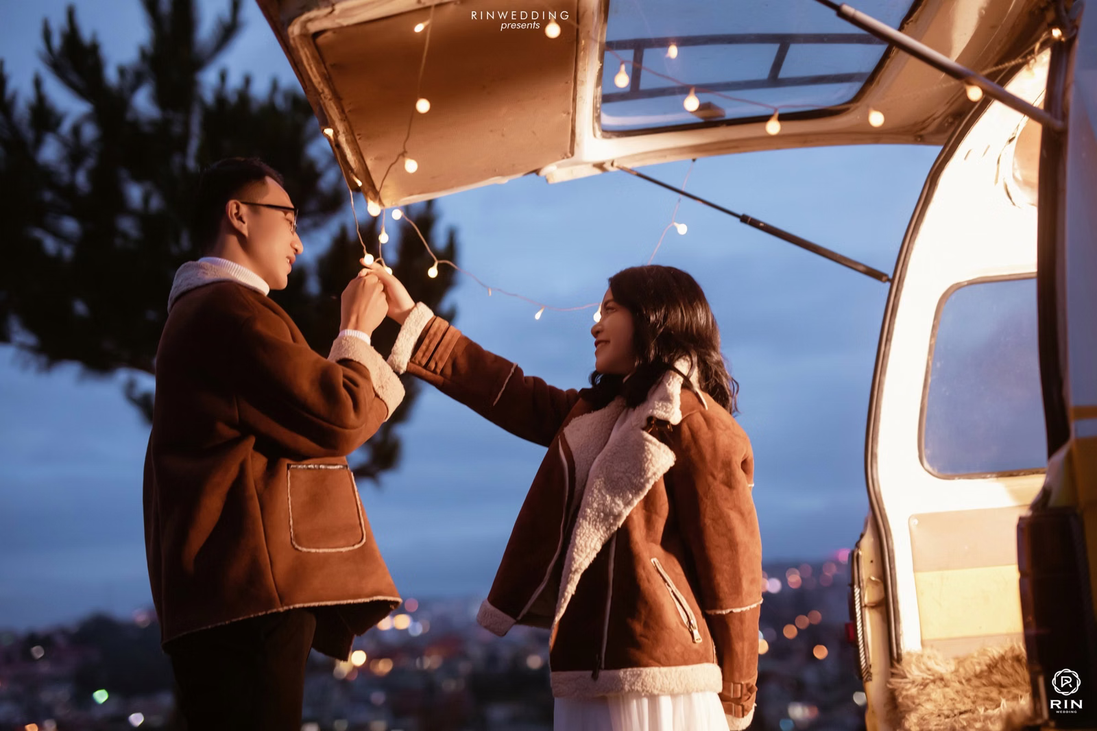 A couple touches hands beneath string lights beside a camper after dark.