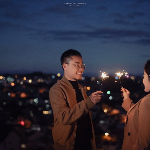 The couple holding sparklers and looking at each other against city lights at night.