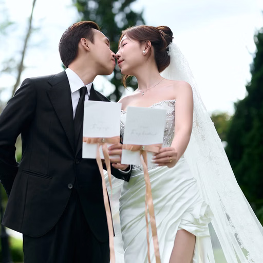 The couple kisses while holding a wedding invitation in a resort garden.