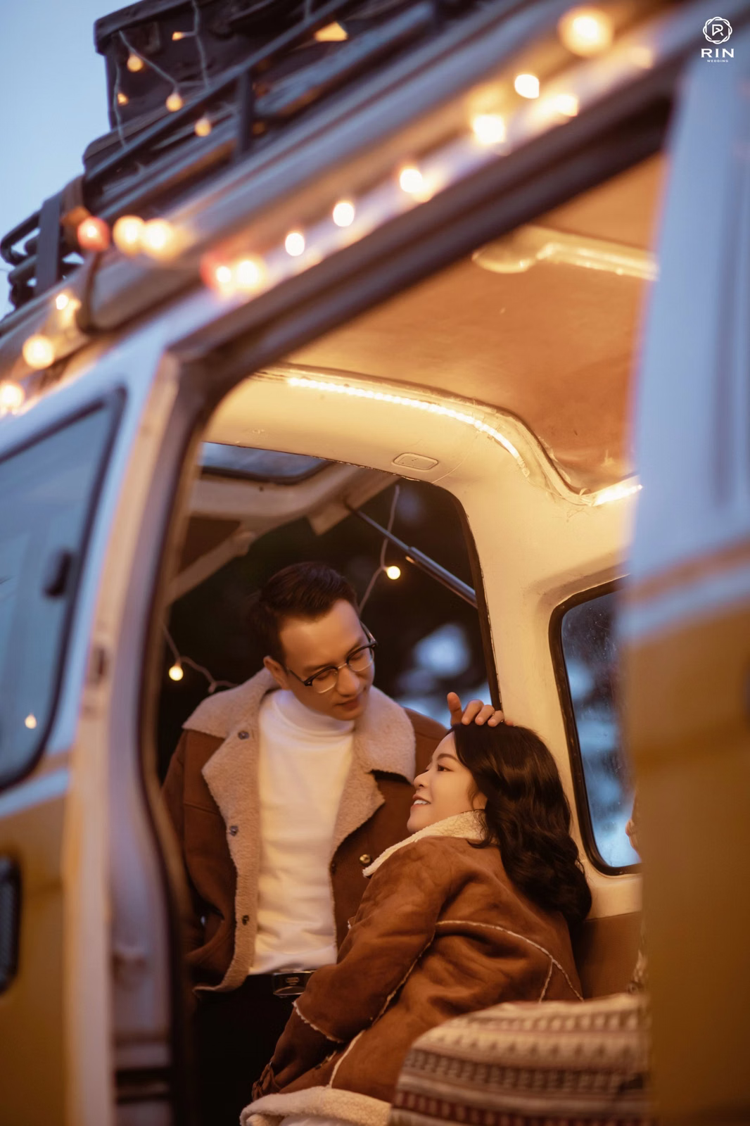 A groom looks at the bride seated in a camper beneath warm lights.