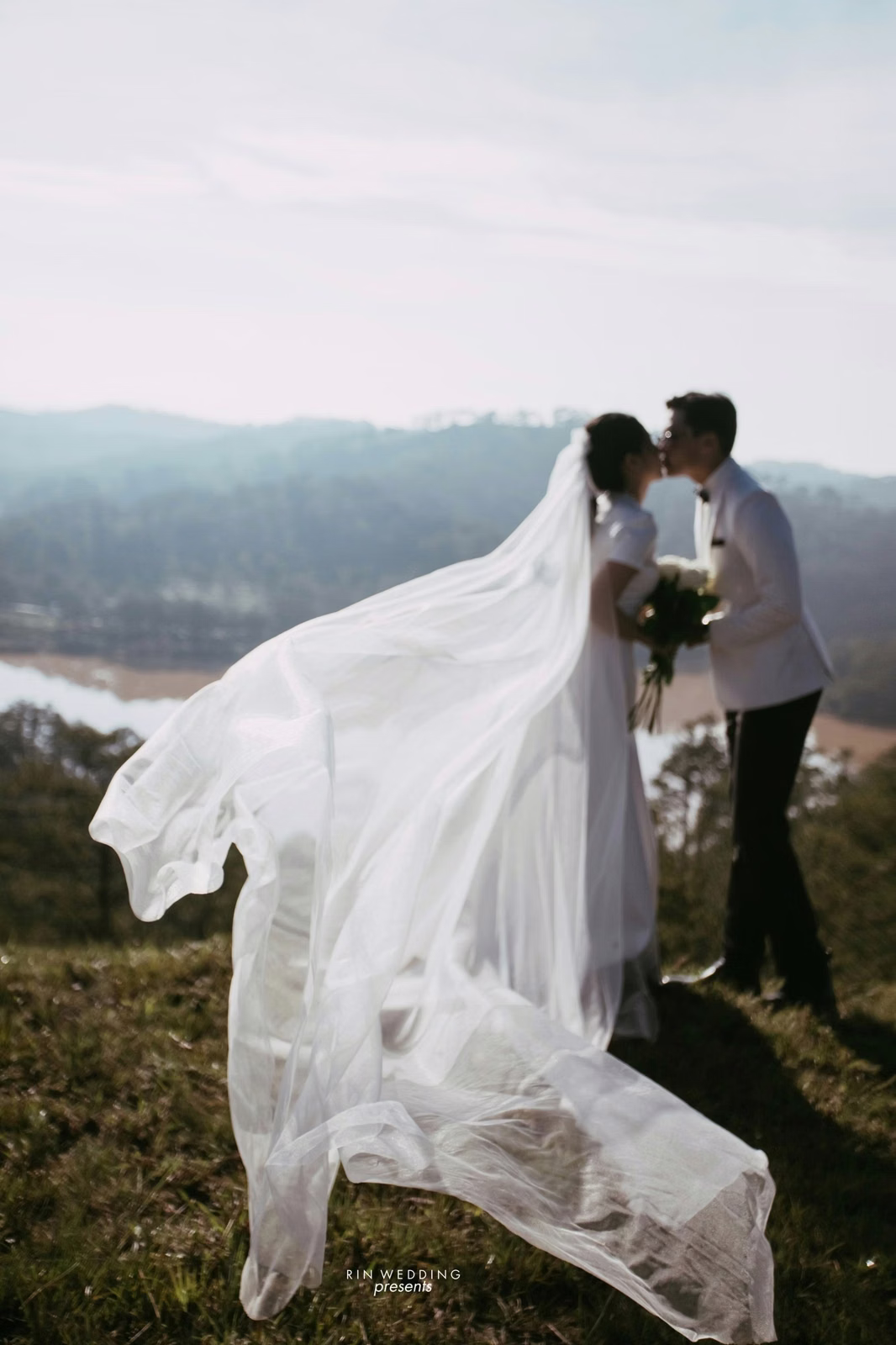 A veil flows behind the bride standing beside the groom at Đồi Thông Đà Lạt