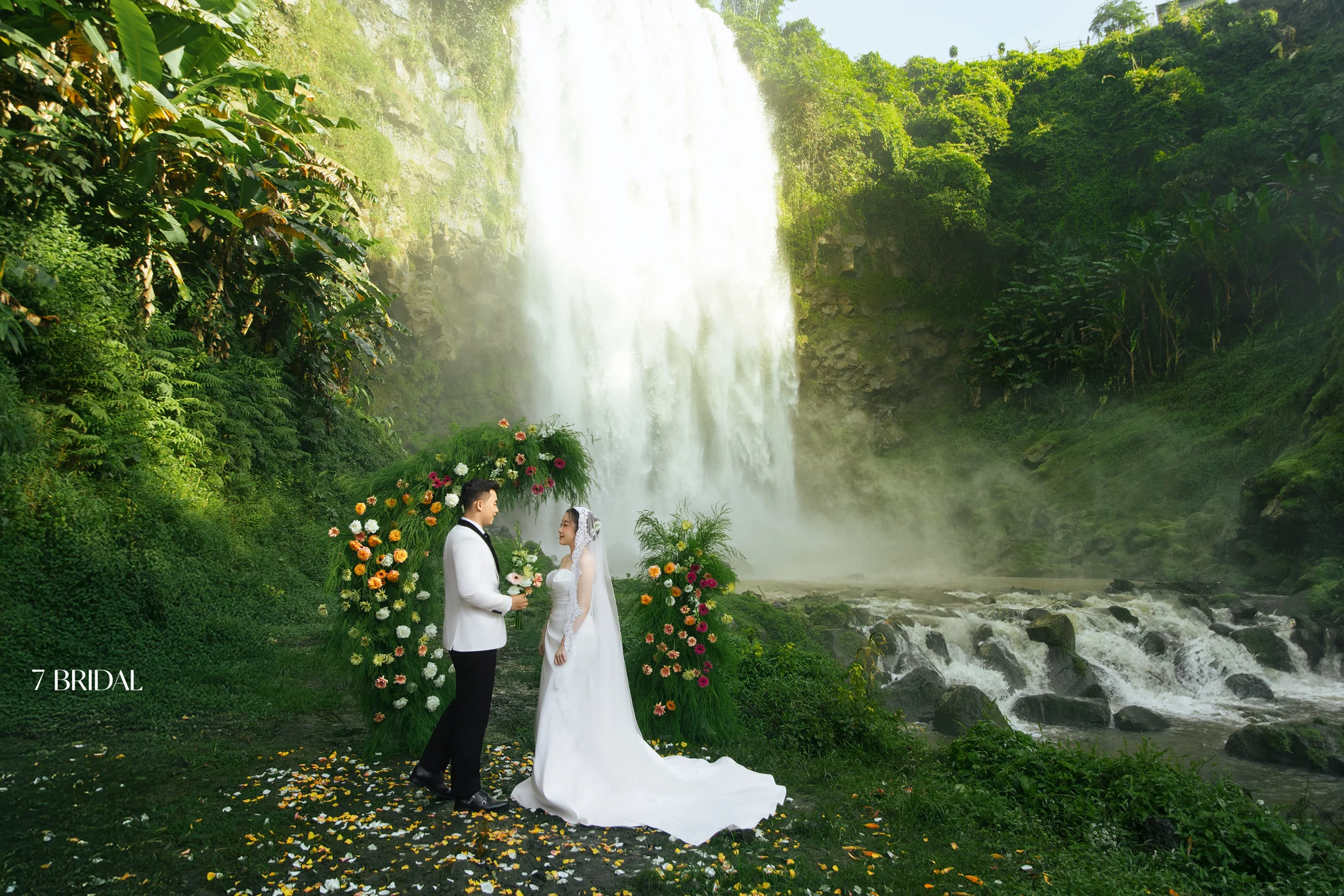 A bride and groom face each other beneath a floral arch before Thác Liêng Chi Nha.