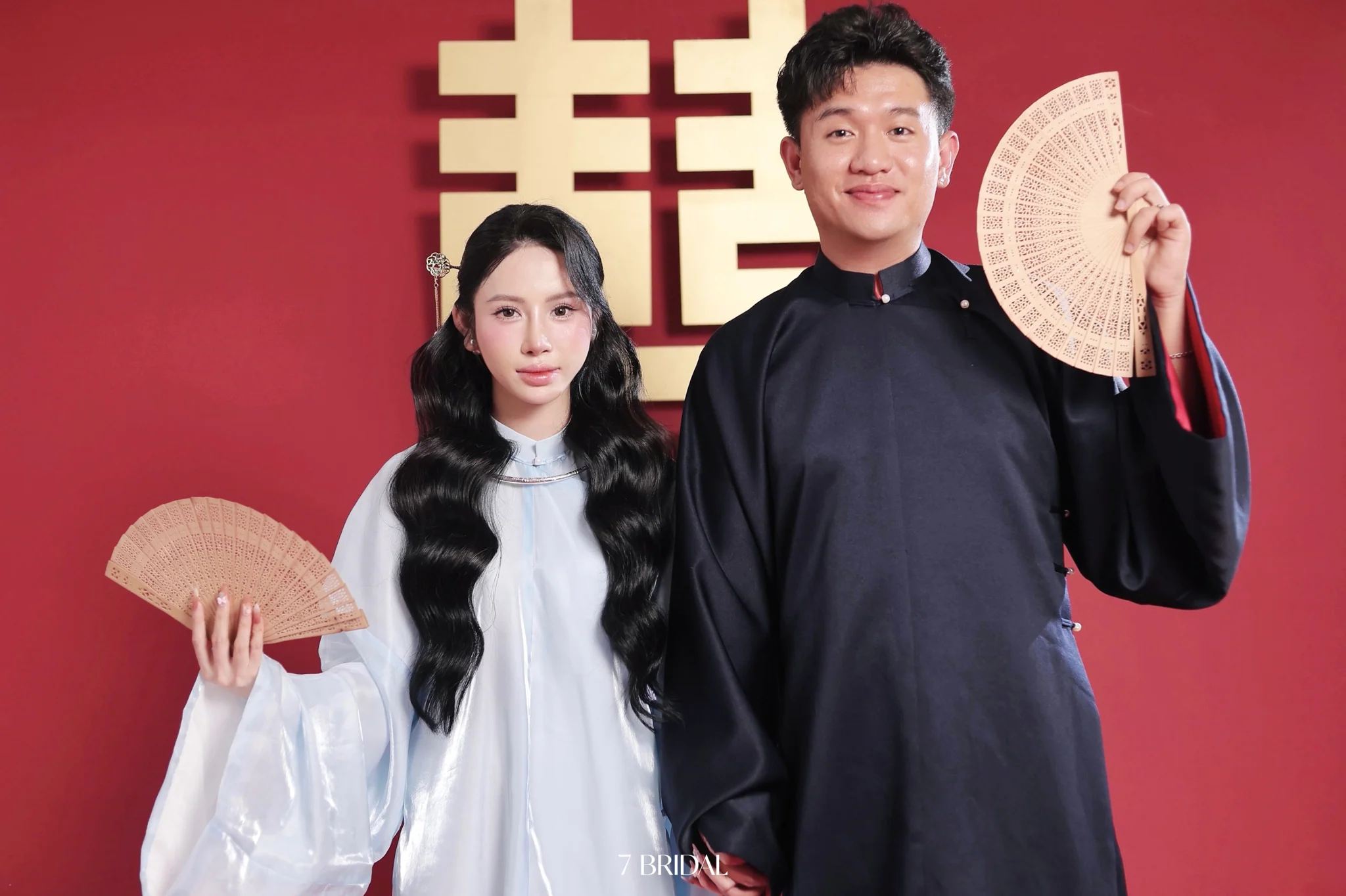 Cặp đôi mặc áo dài và cầm quạt trước chữ hỷ màu vàng.